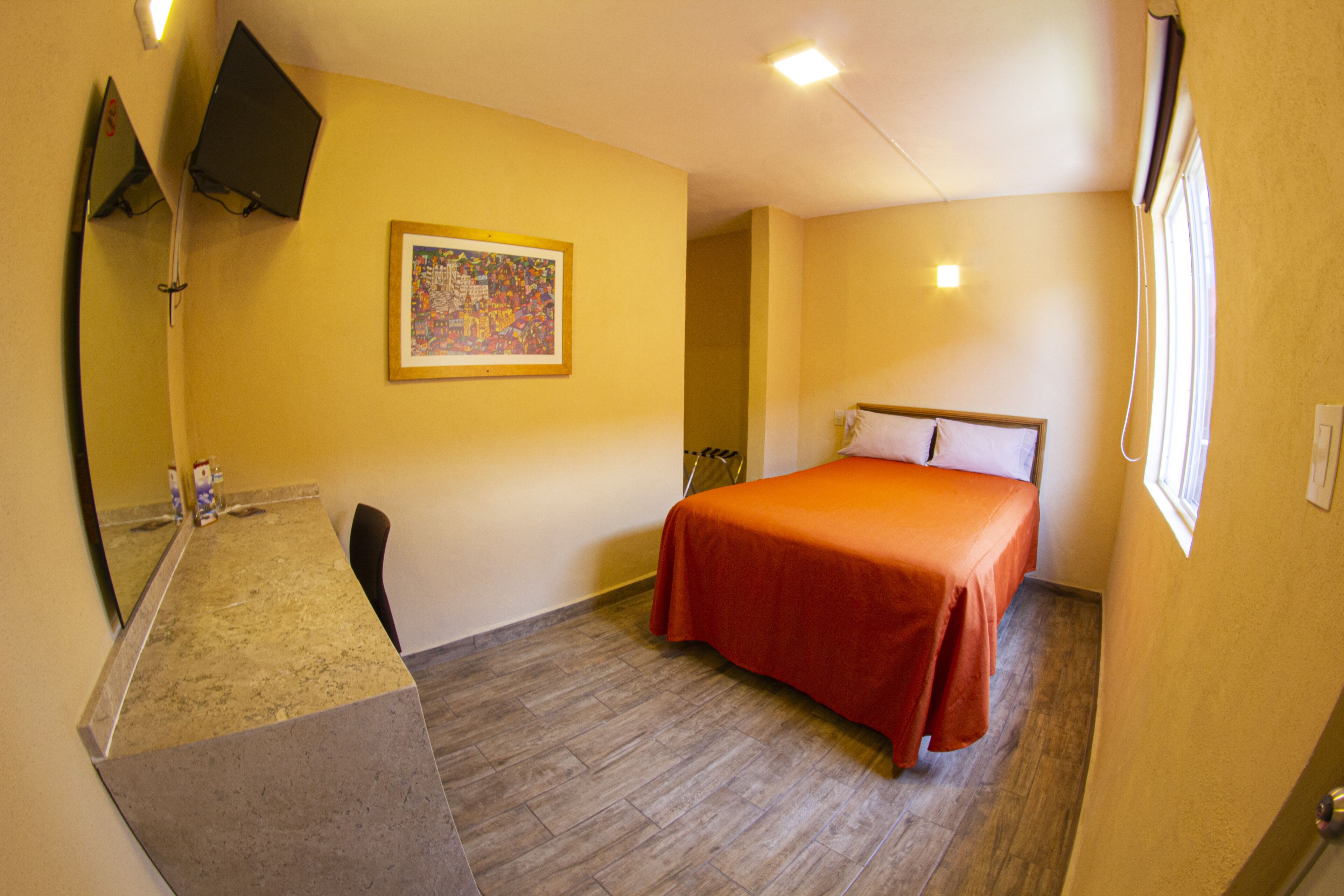 A hotel room with a bed, a desk, a chair, a mirror, a painting on the wall, and a window.