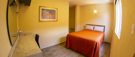 A hotel room with a bed, a desk, a chair, a mirror, a painting on the wall, and a window.