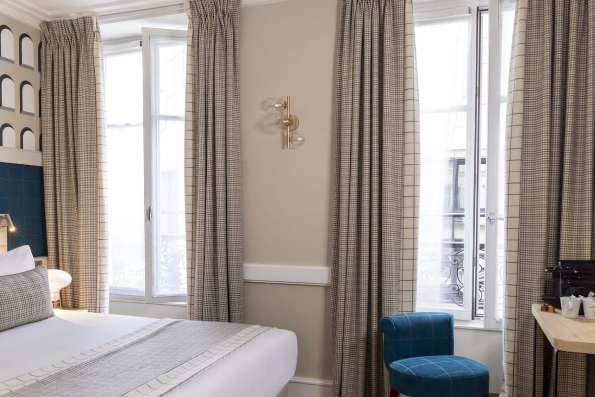 Hotelkamer met een groot bed, geruite gordijnen, een klein tafeltje met een plantje en uitzicht buiten.