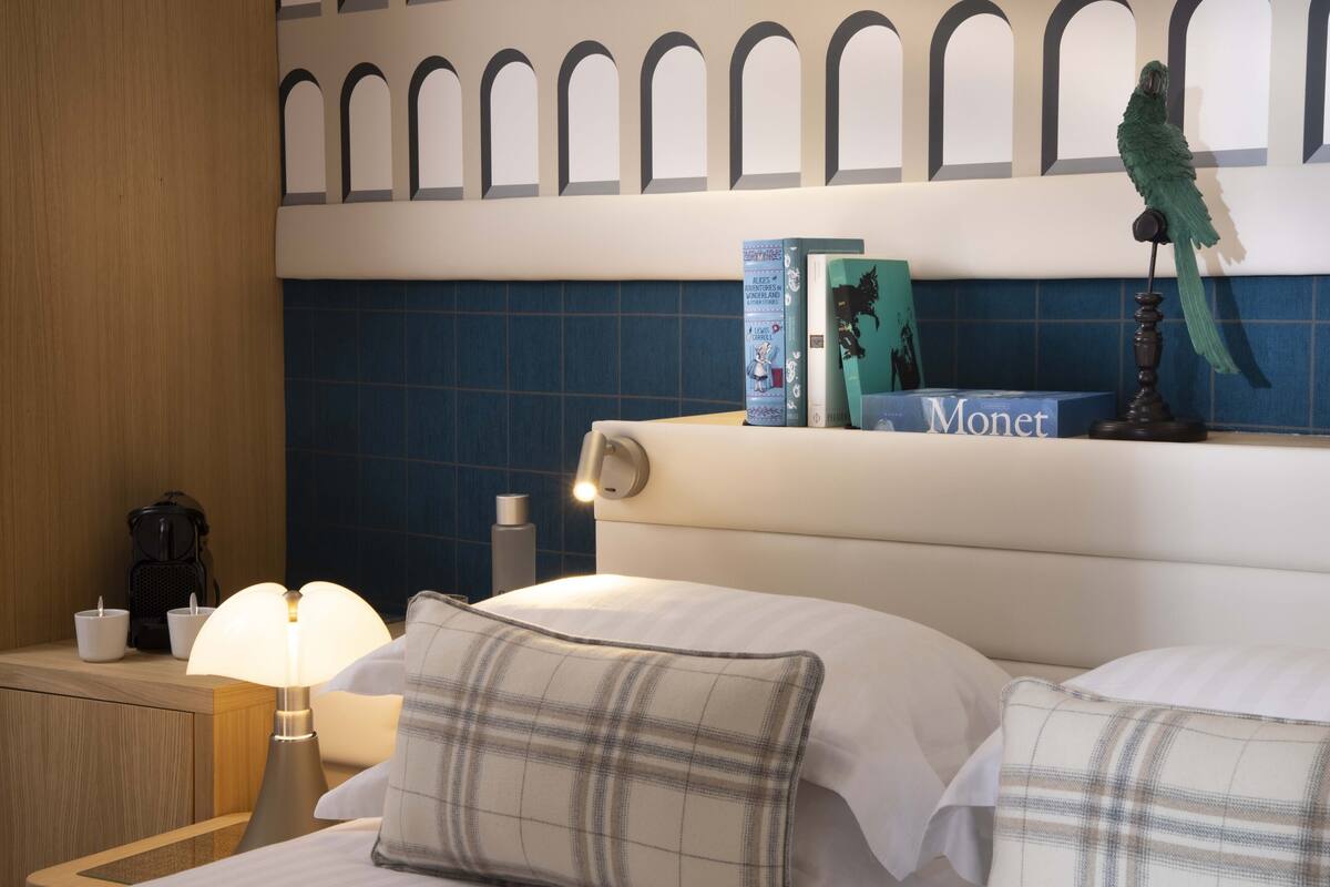Een hotelkamer met een bed, een nachtkastje met een lamp, een koffiezetapparaat en boeken op het hoofdeinde.