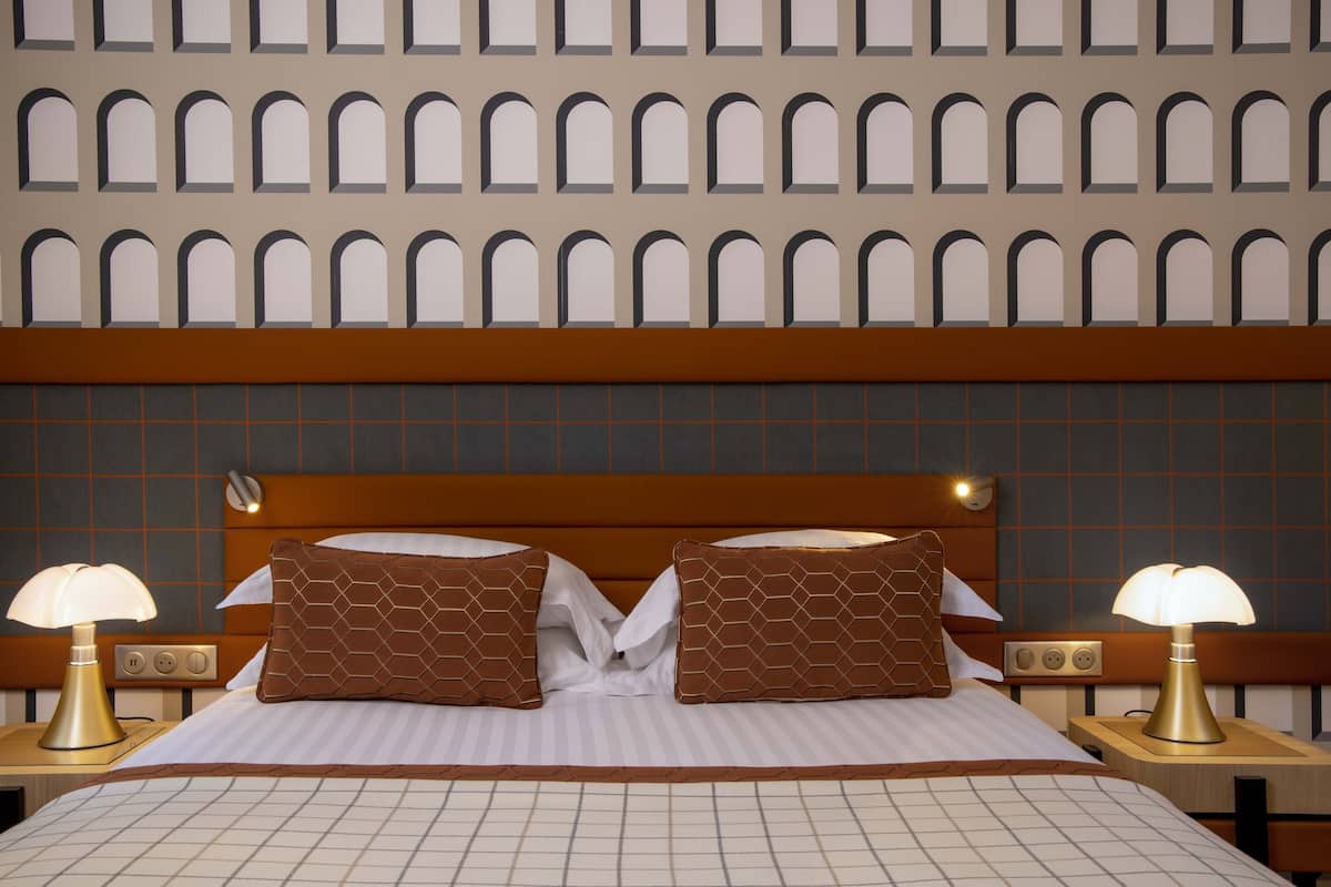 Een hotelkamer met een groot bed, twee nachtkastjes, een muur met boogvensters en een hoofdbord met een rasterpatroon.