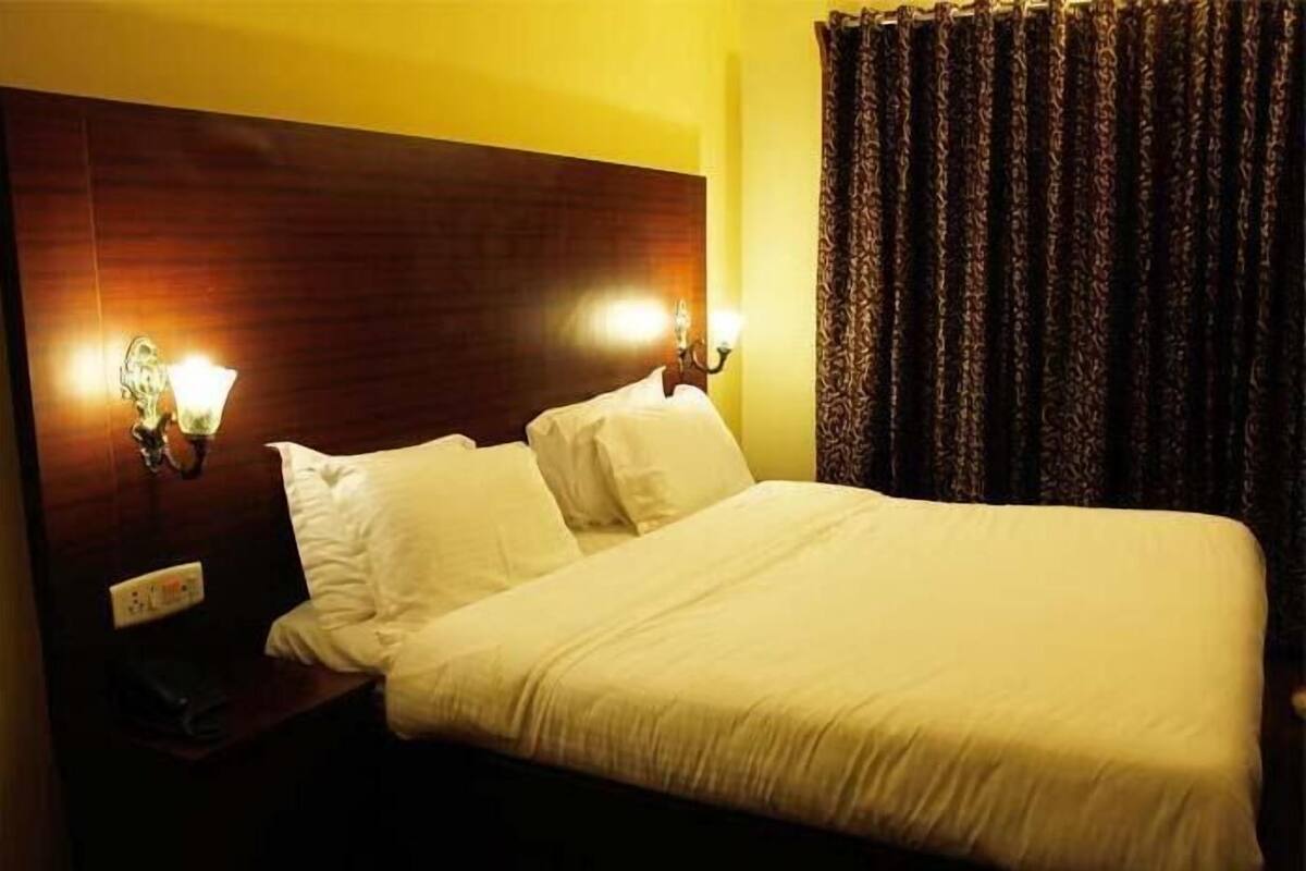 A hotel room with a wooden headboard, two bedside lamps, a bed with white linens, and a dark patterned curtain.