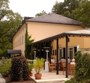 An outdoor cafe with tables and chairs, surrounded by greenery and a building with a tiled roof. - Landhaus Lockwitzgrund (Dresden)