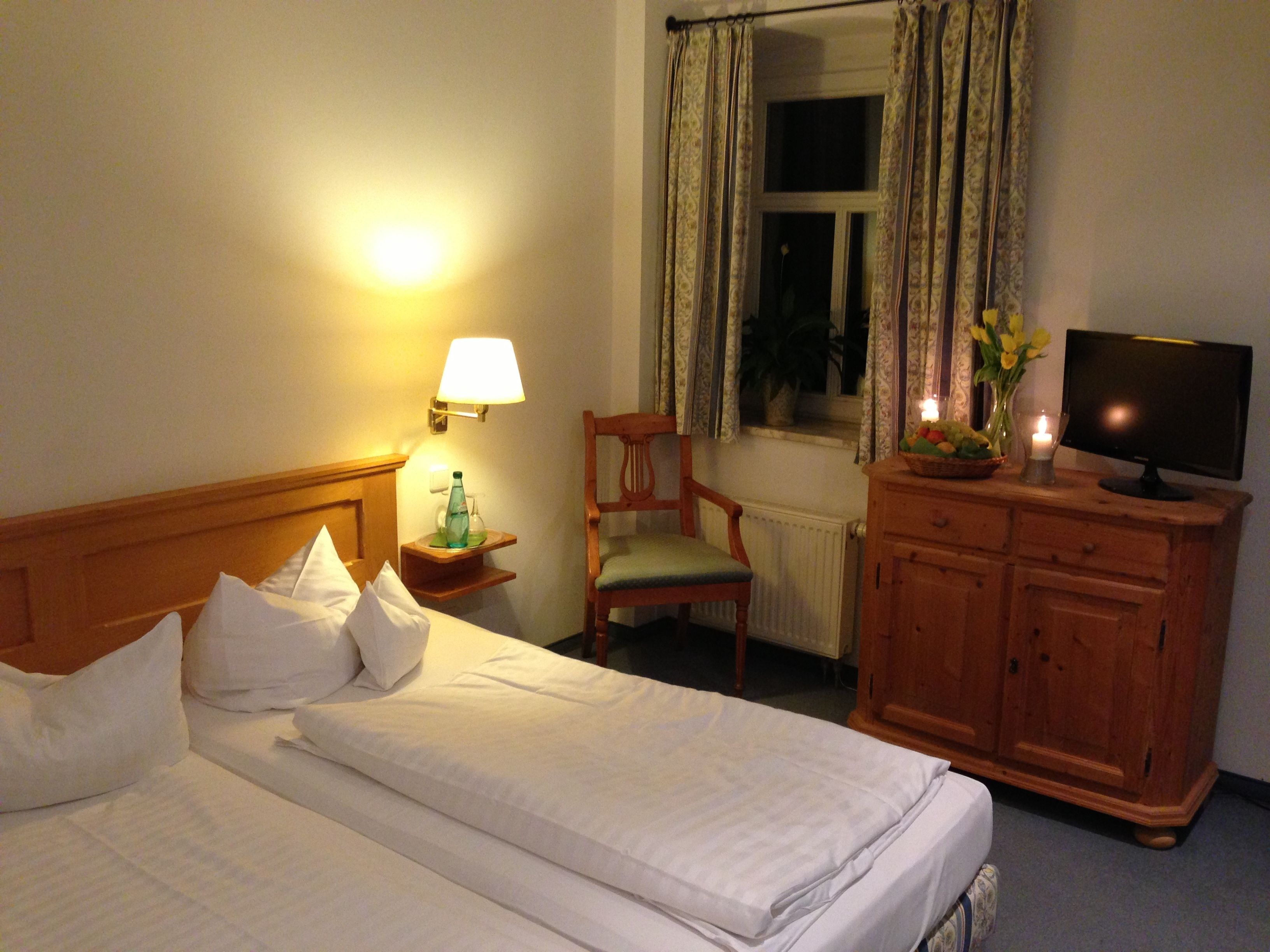 A hotel room with a neatly made bed, a wooden nightstand with a lamp, a chair, a television on a wooden cabinet, and a window with curtains.