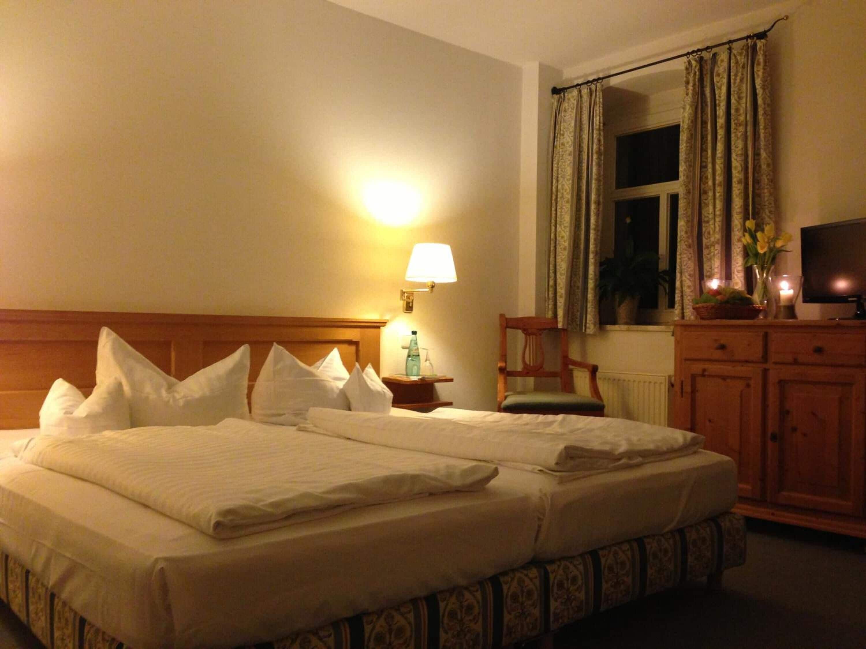 A hotel room with a large bed, a nightstand with a lamp, a wooden chair, a television, and a window with curtains.