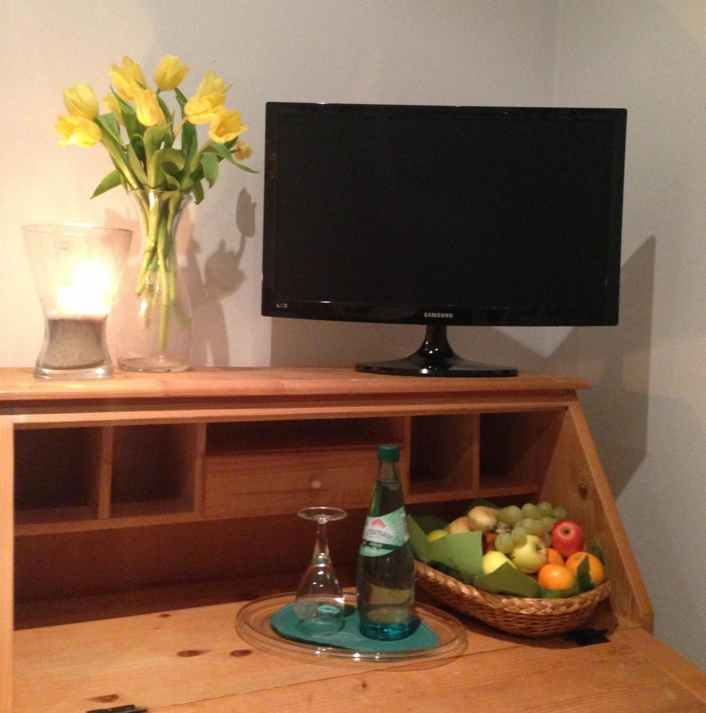 A flat-screen TV on a wooden stand, a vase with yellow flowers, a candle, a bottle of water, and a basket of fruit.