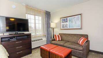 Habitación de hotel con televisión de pantalla plana, un sofá, un reposapiés, cómoda y un cuadro en la pared.