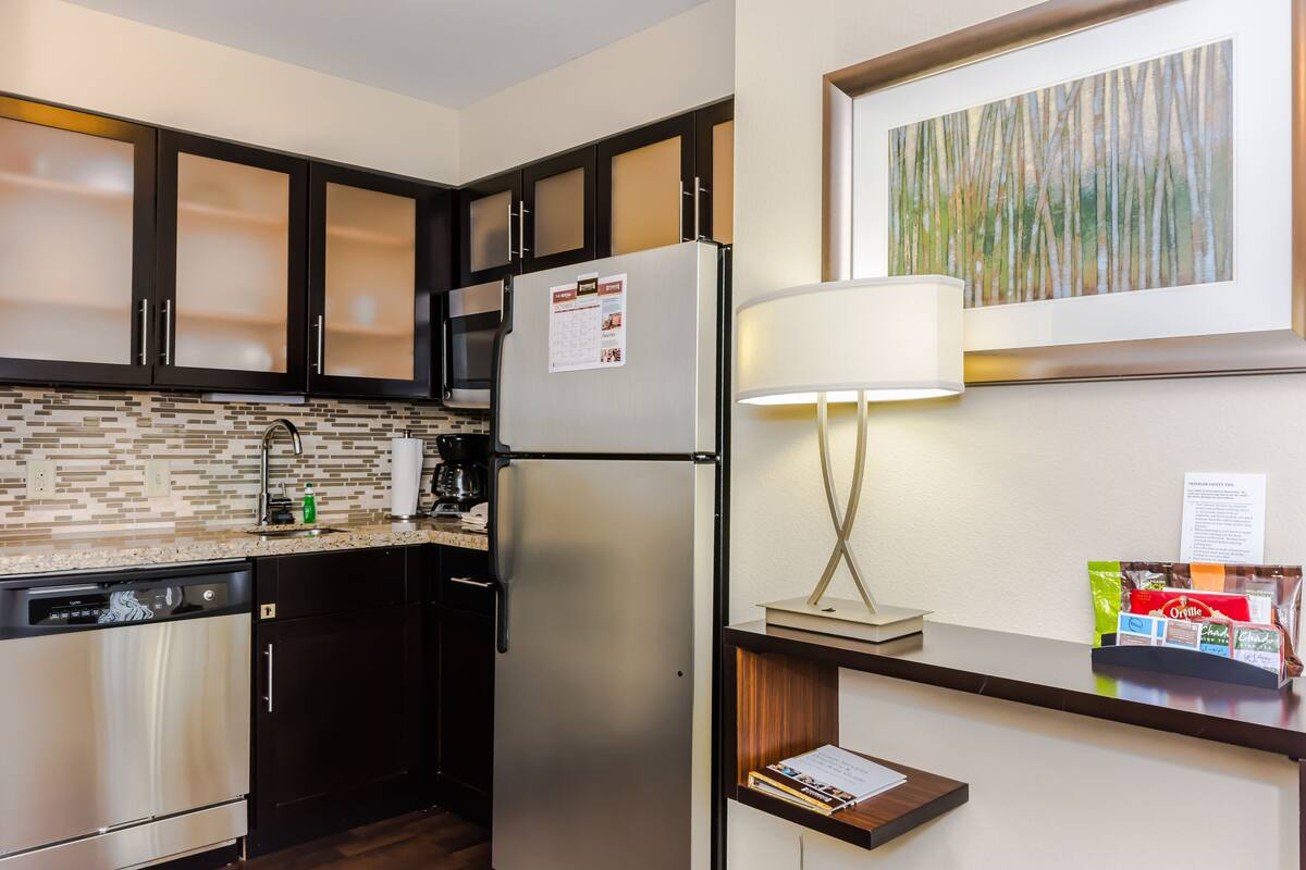 A compact kitchen with dark cabinets, a stainless steel refrigerator, a coffee maker, and a small shelf with a lamp and a framed picture.