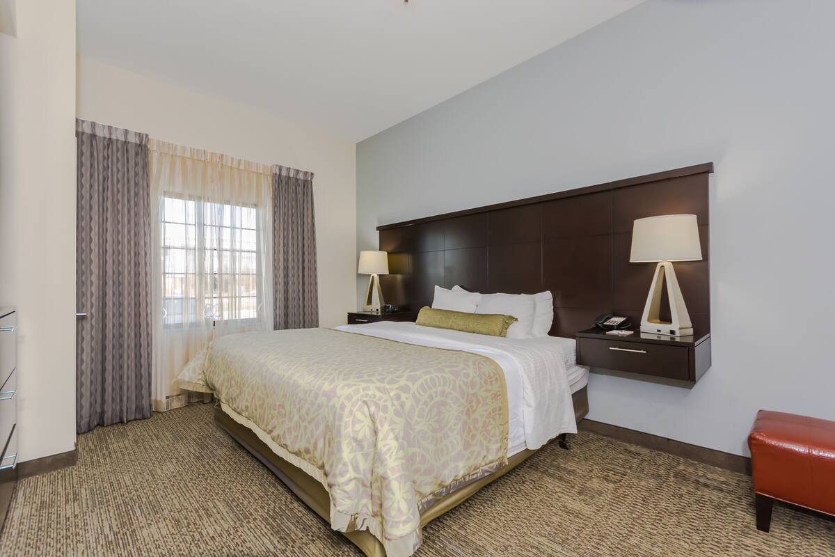 A hotel room with a large bed, a nightstand with a lamp, a bedside table with a lamp, and a red bench.