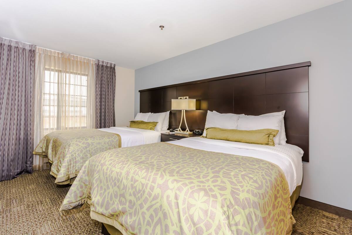 A hotel room with two beds, a nightstand with a lamp, and a large window with curtains.