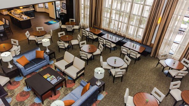 A hotel lobby with multiple seating areas, including sofas, chairs, and tables. There is a counter with a menu and a small bar area. Large windows with curtains provide natural light. - Staybridge Suites Grand Forks by IHG (Grand Forks)