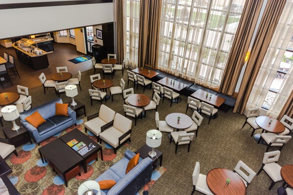 Een hotel lobby met verschillende zithoeken, waaronder banken, stoelen en tafels. Er is een balie met een menukaart en een klein barretje. Grote ramen met gordijnen zorgen voor natuurlijk licht.