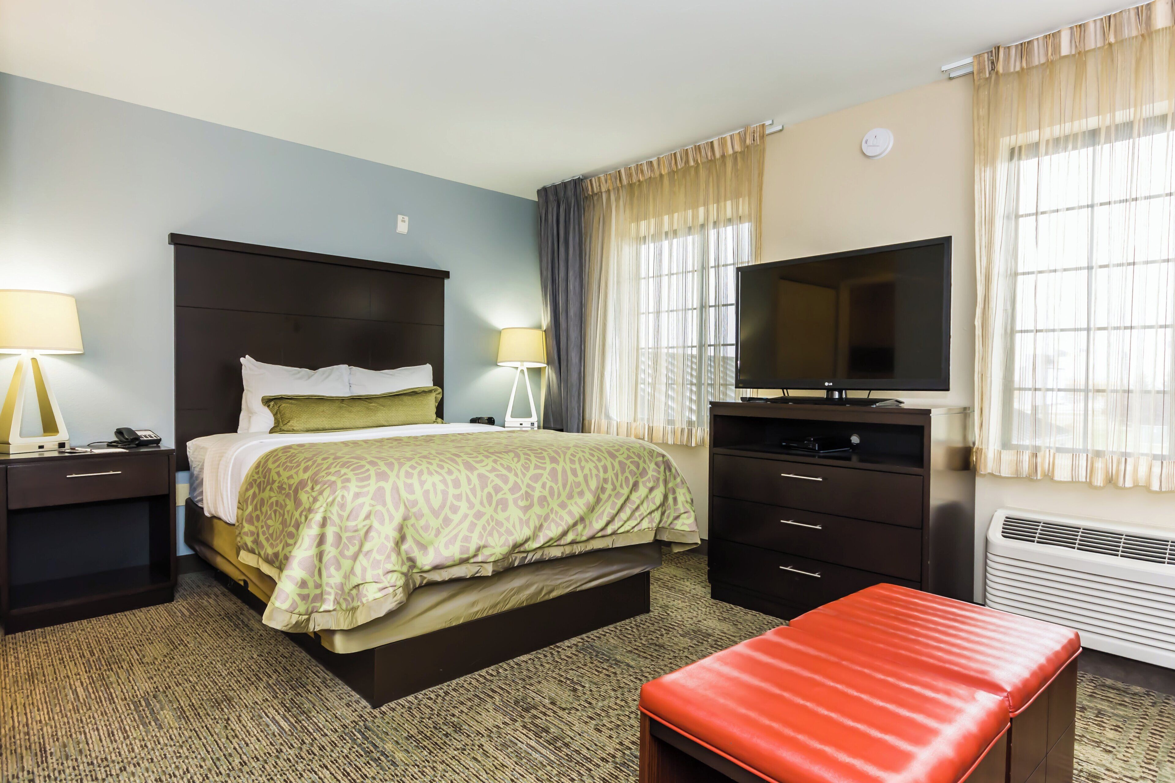 A hotel room with a bed, bedside tables, a TV, a desk, and a red bench.