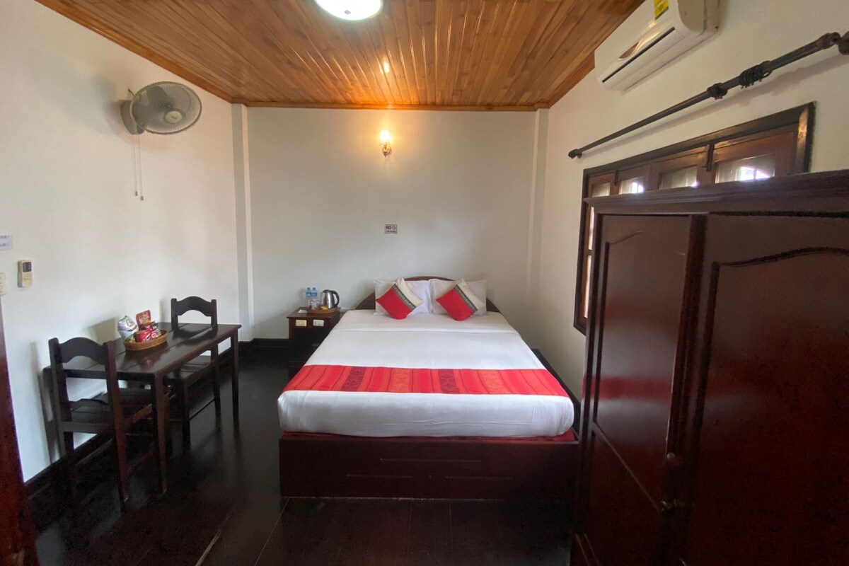 A hotel room with a wooden ceiling, a bed with a red and white bedspread, a desk with chairs, a nightstand with a kettle, and a wardrobe.
