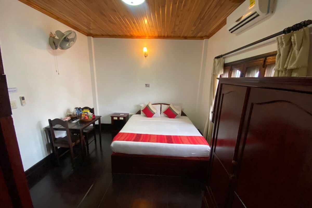 A hotel room with a wooden ceiling, a bed with red and white bedding, a desk with a chair, a wall-mounted fan, and a wardrobe.