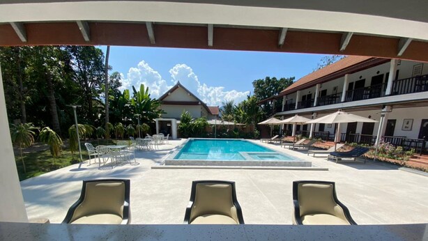 A pool area with lounge chairs, a building with a red roof, and a clear sky. - Vangsavath Hotel (Luang Prabang)