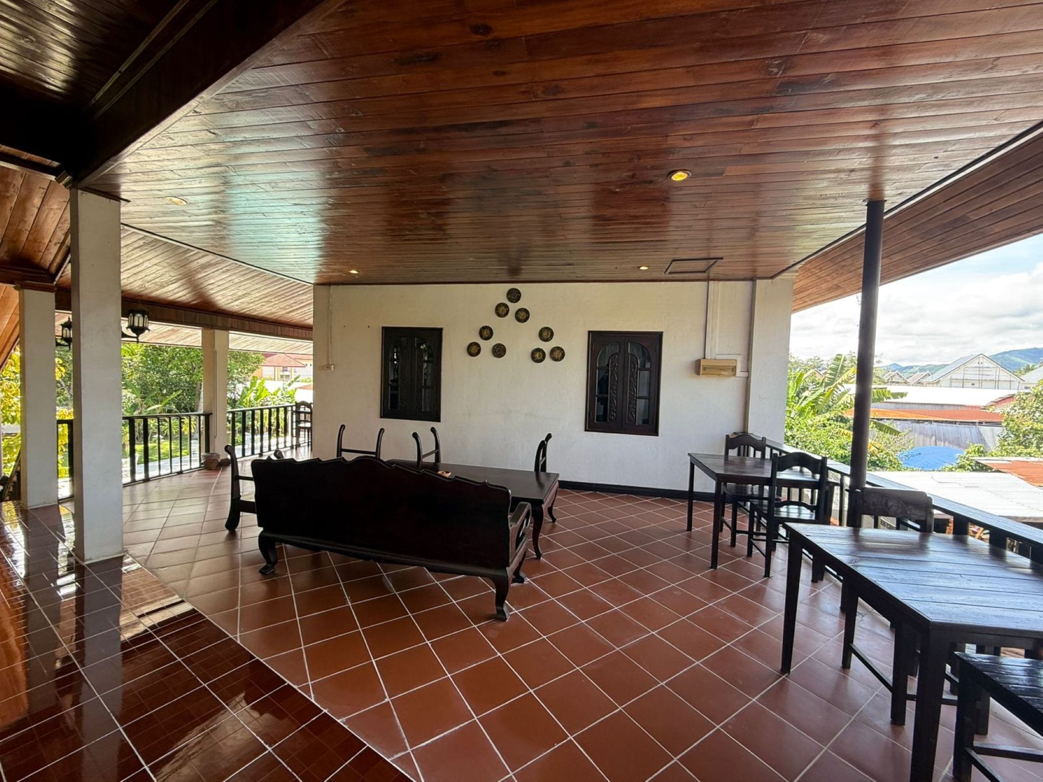 A covered patio with a black sofa, dining tables, and chairs, featuring a tiled floor and a wooden ceiling.