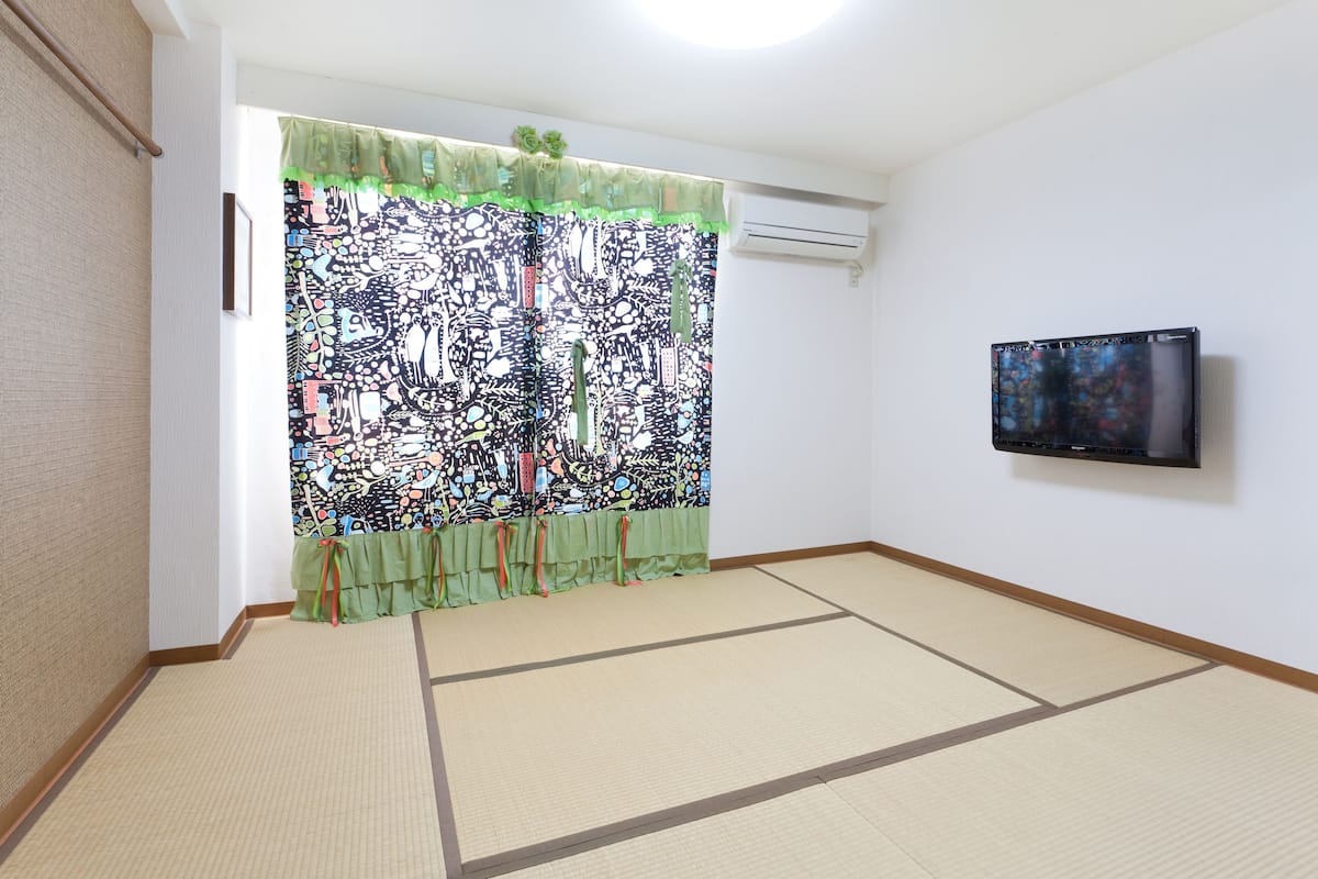 A room with tatami flooring, a green curtain with a colorful pattern, and a flat-screen TV mounted on the wall.