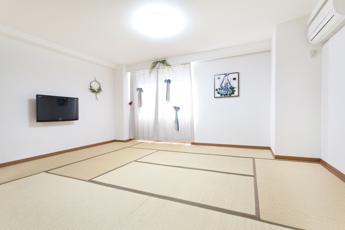 A room with tatami flooring, a mounted TV, a framed picture, and a window with sheer curtains.