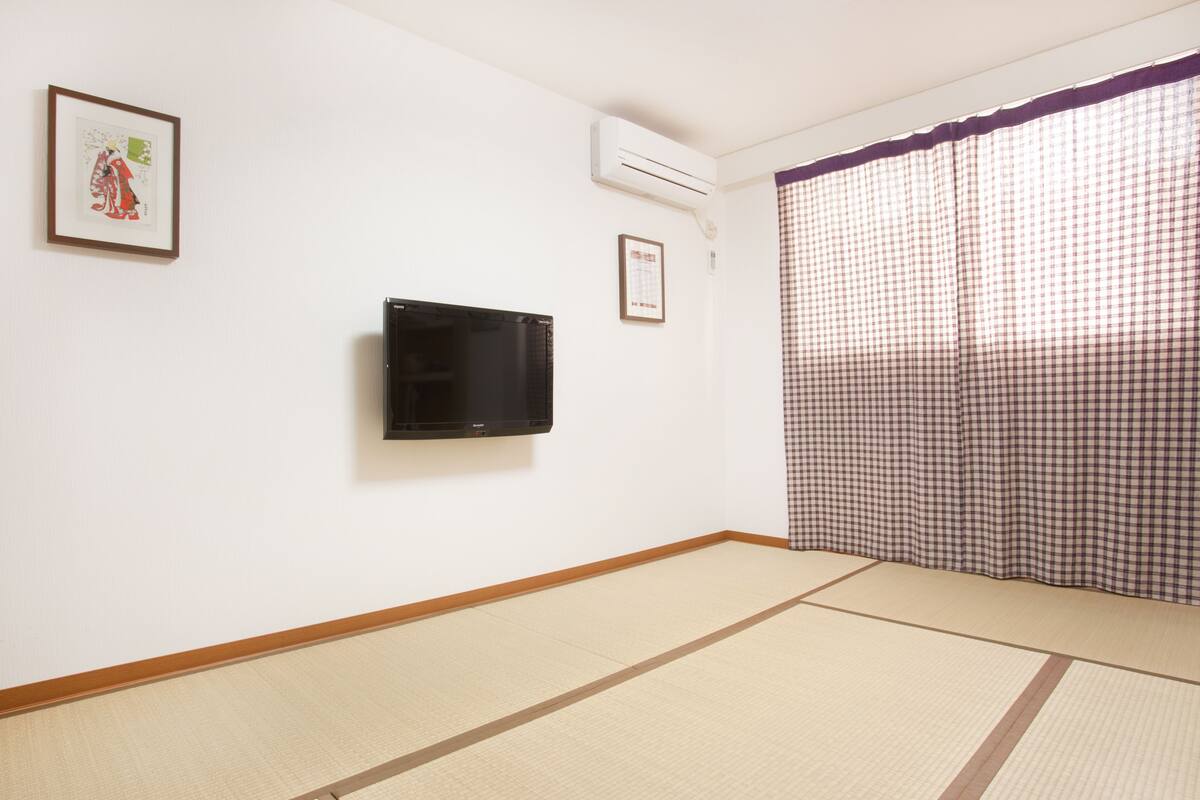 A room with tatami flooring, a mounted TV, an air conditioning unit, and a checkered curtain.