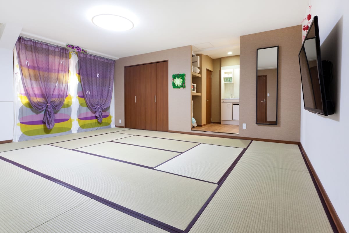 A room with tatami flooring, a sliding door with purple curtains, a wooden wardrobe, a mirror, and a television.