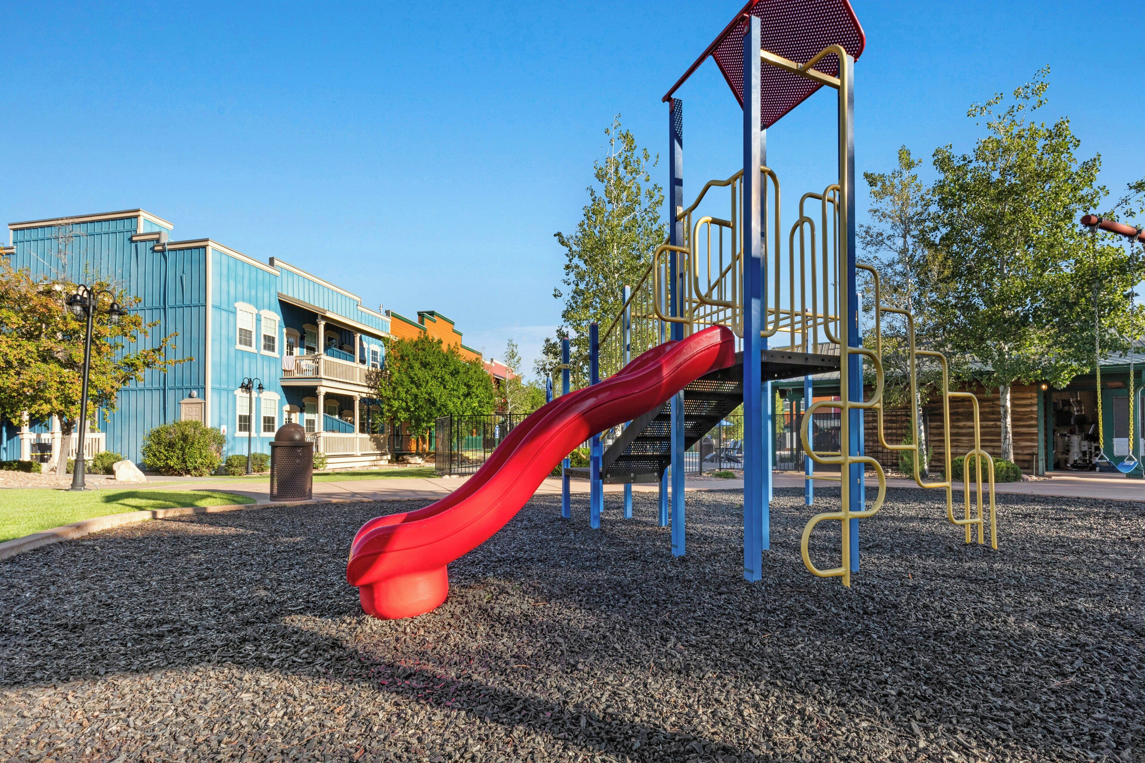 A playground with a red slide and climbing structures, surrounded by a gravel area and buildings in the background.