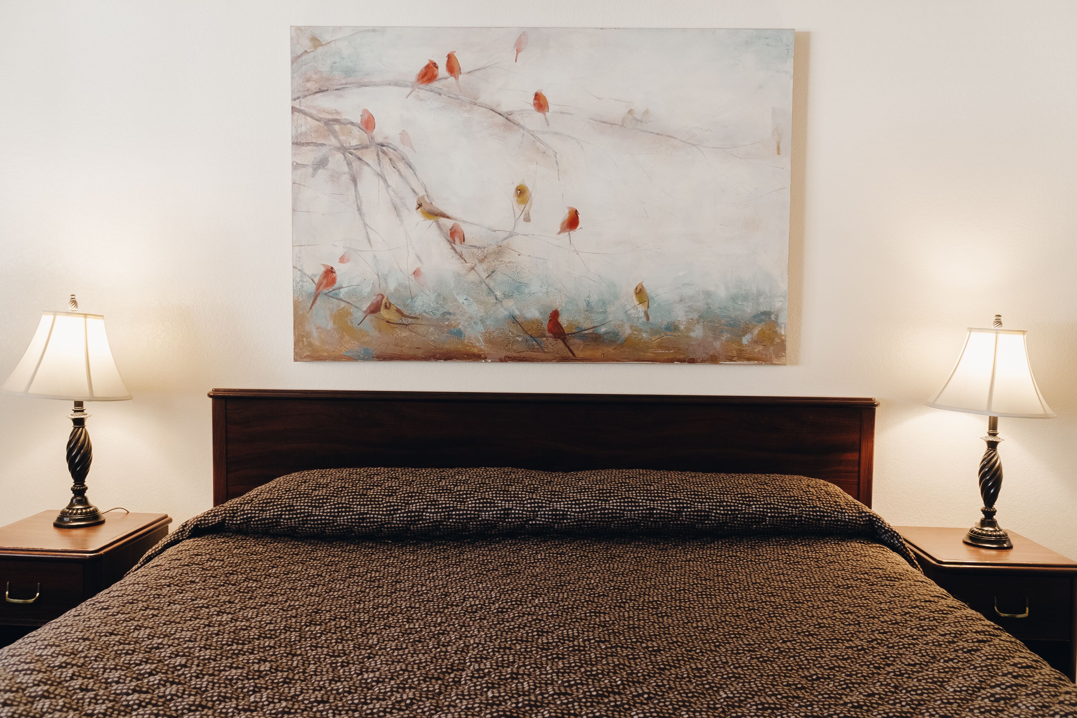 Un letto rifatto con cura, con una coperta marrone a trama decorativa, affiancato da due comodini con lampade, e un quadro astratto appeso alla parete sopra.