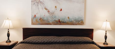 Una cama bien tendida con una manta marrón texturizada, flanqueada por dos mesitas de noche con lámparas, y un cuadro abstracto colgado en la pared.
