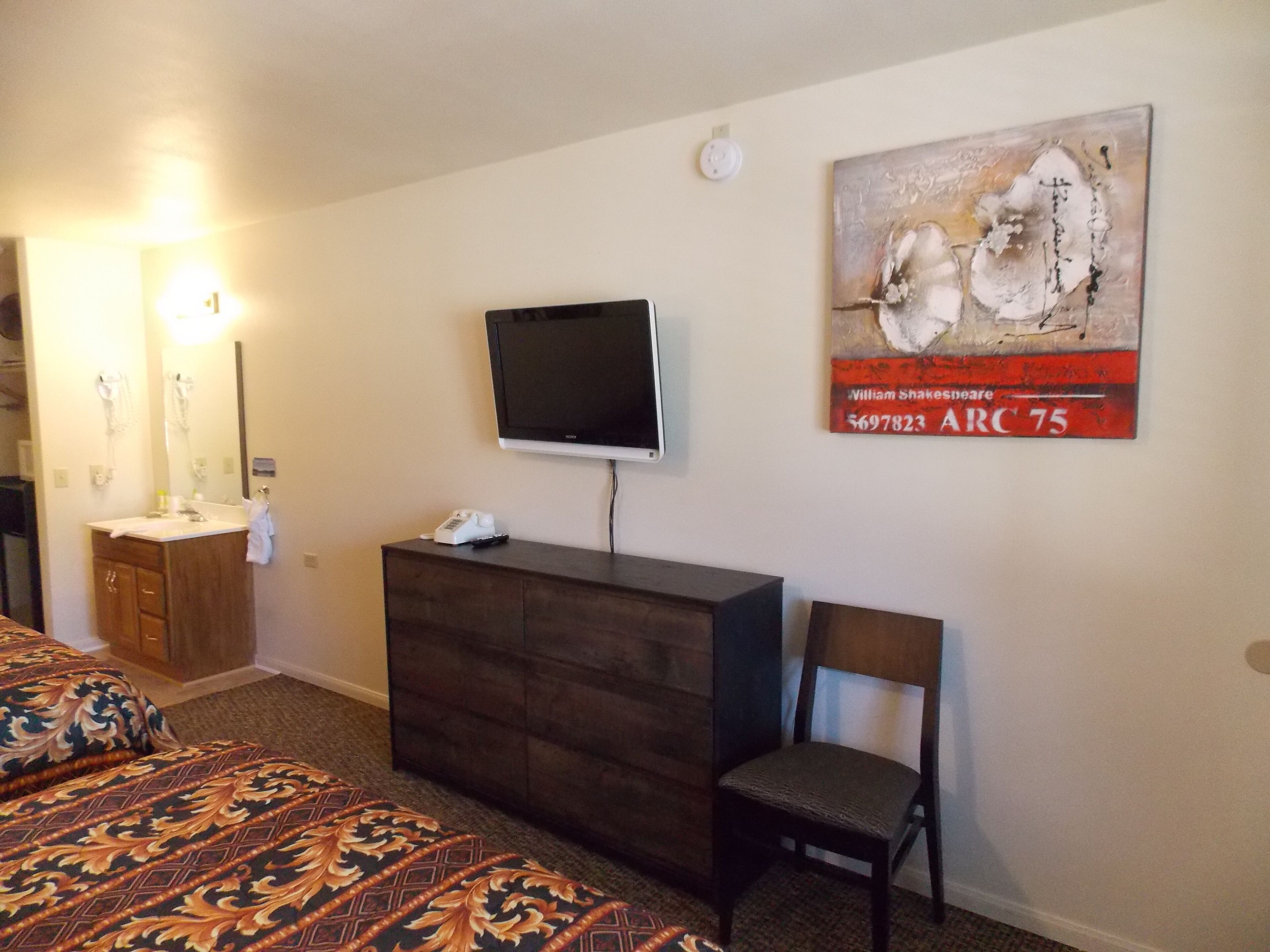 A hotel room with a bed, a dresser, a television mounted on the wall, a chair, and a painting.