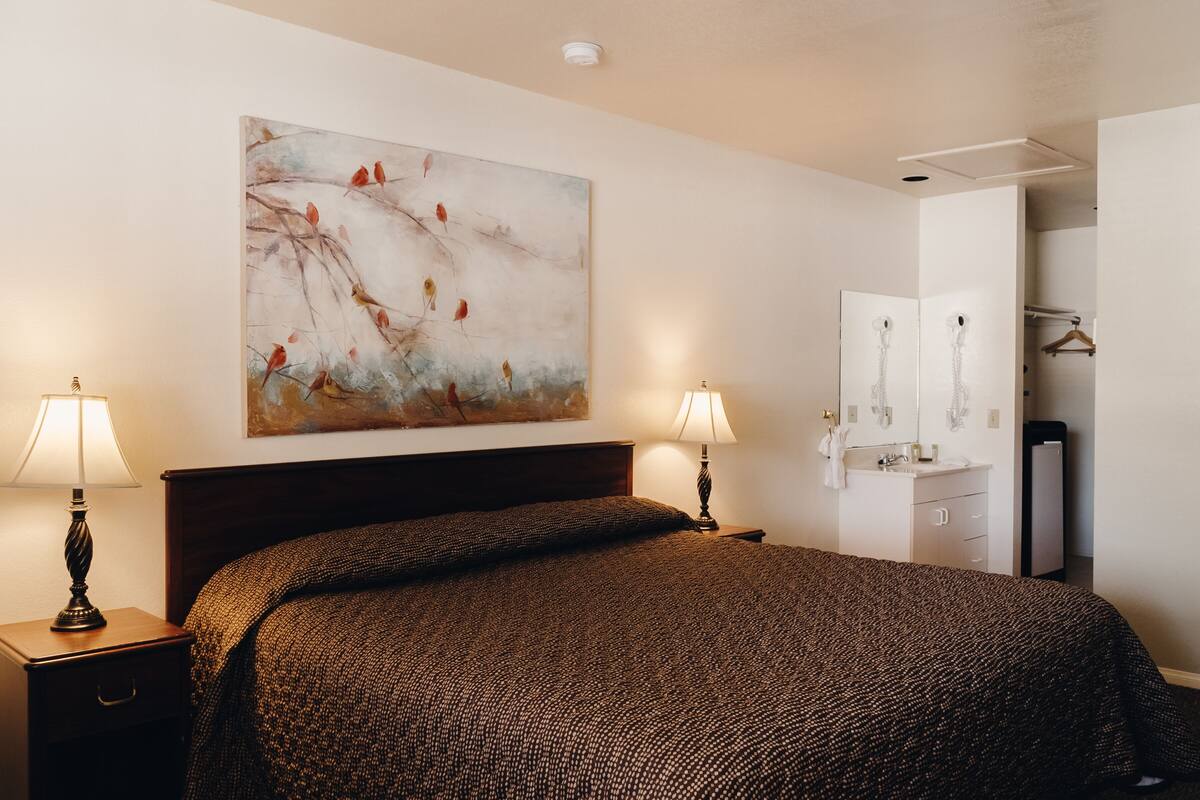 A neatly made bed with a patterned bedspread, two bedside lamps, a wall-mounted painting of birds, and a partially visible bathroom with a mirror and towel rack.