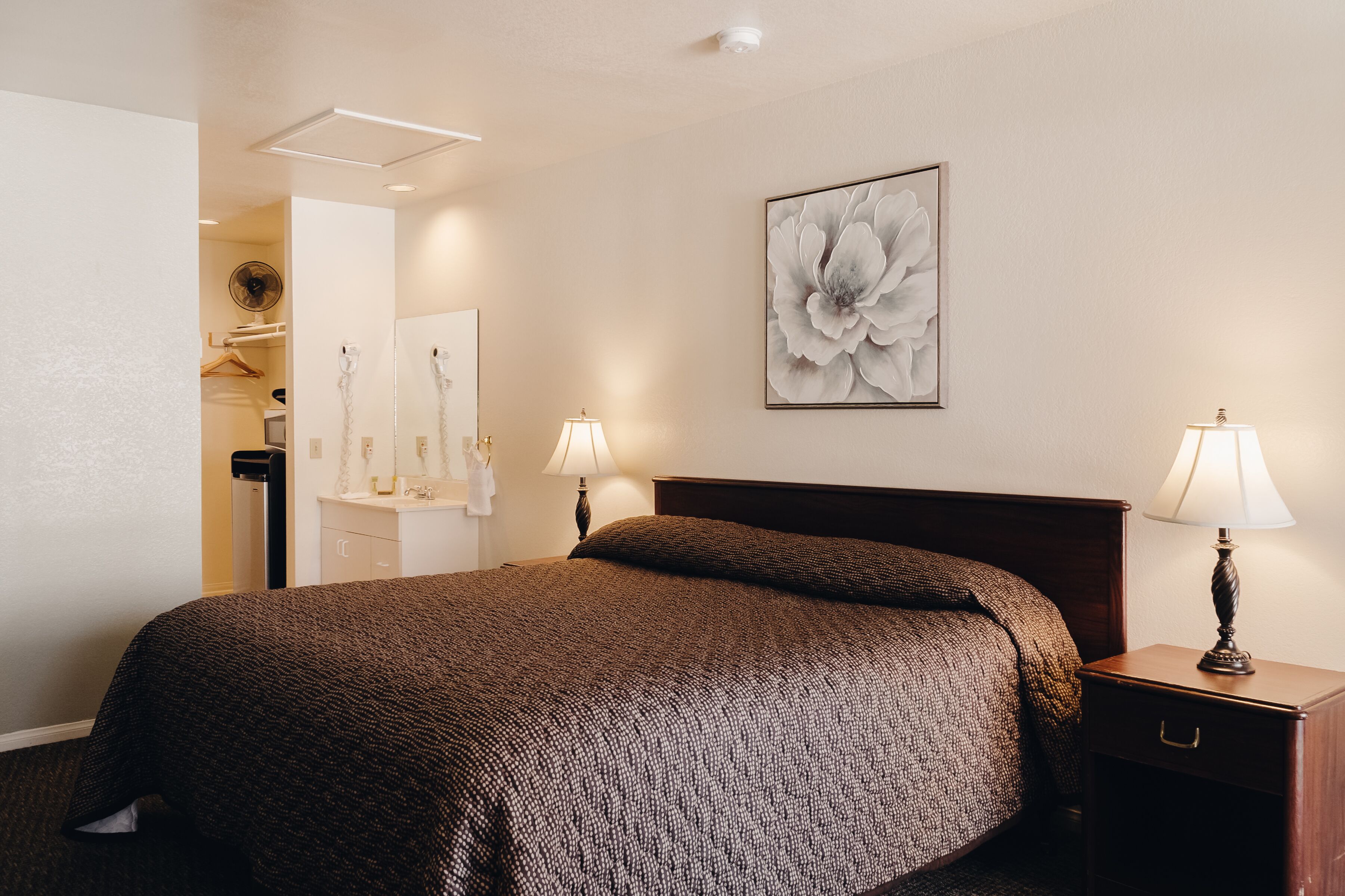 A neatly made bed with a dark patterned bedspread, two bedside lamps, and a large floral artwork on the wall.