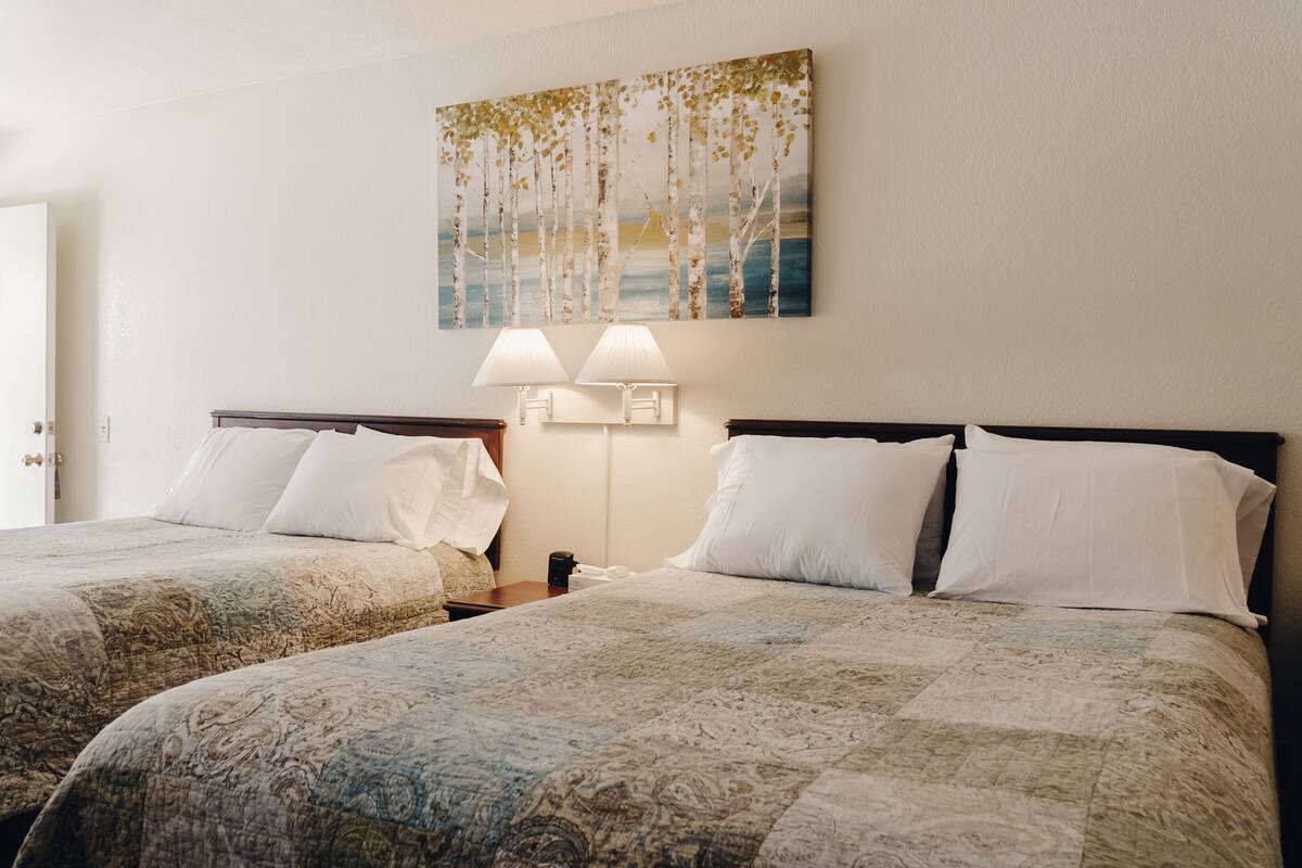 A hotel room with two beds, a painting of trees, and two wall-mounted lamps.