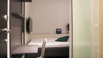 A compact room with a bed, a small table, a chair, a mounted TV, and a potted plant.