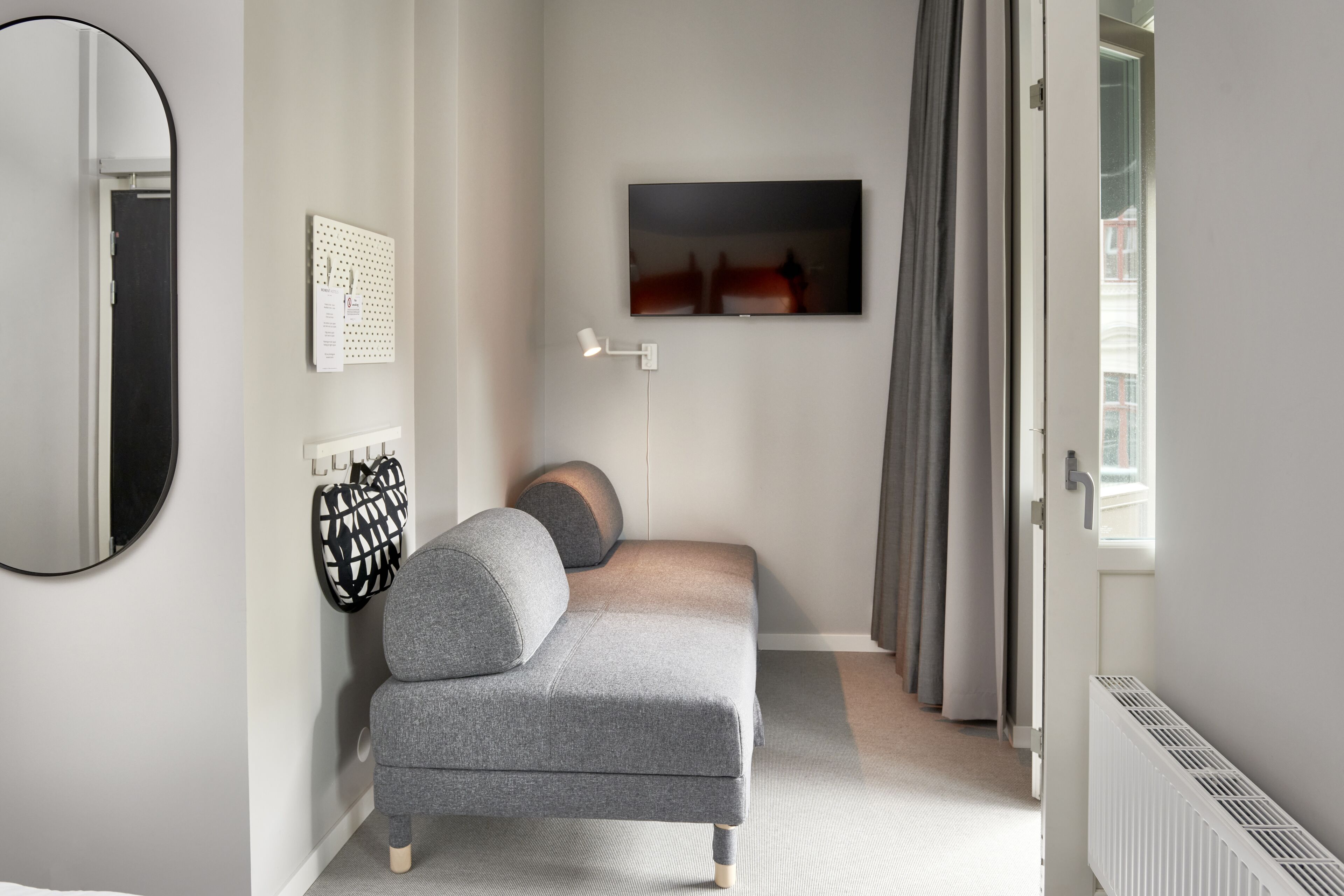 A modern bedroom with a grey sofa, a wall-mounted TV, a mirror, and a radiator.