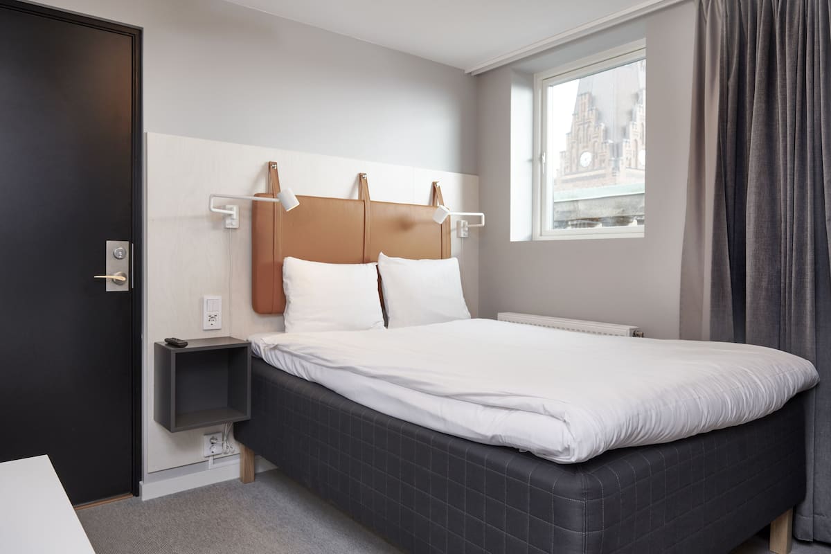 A modern hotel room with a large bed, a bedside table, a nightstand, a window with a view of a building, and a door.