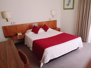 Een hotelkamer met een houten hoofdeinde, twee nachtkastjes met lampen, een telefoon, een rood-witte dekbedovertrek en een ingelijst schilderij aan de muur.