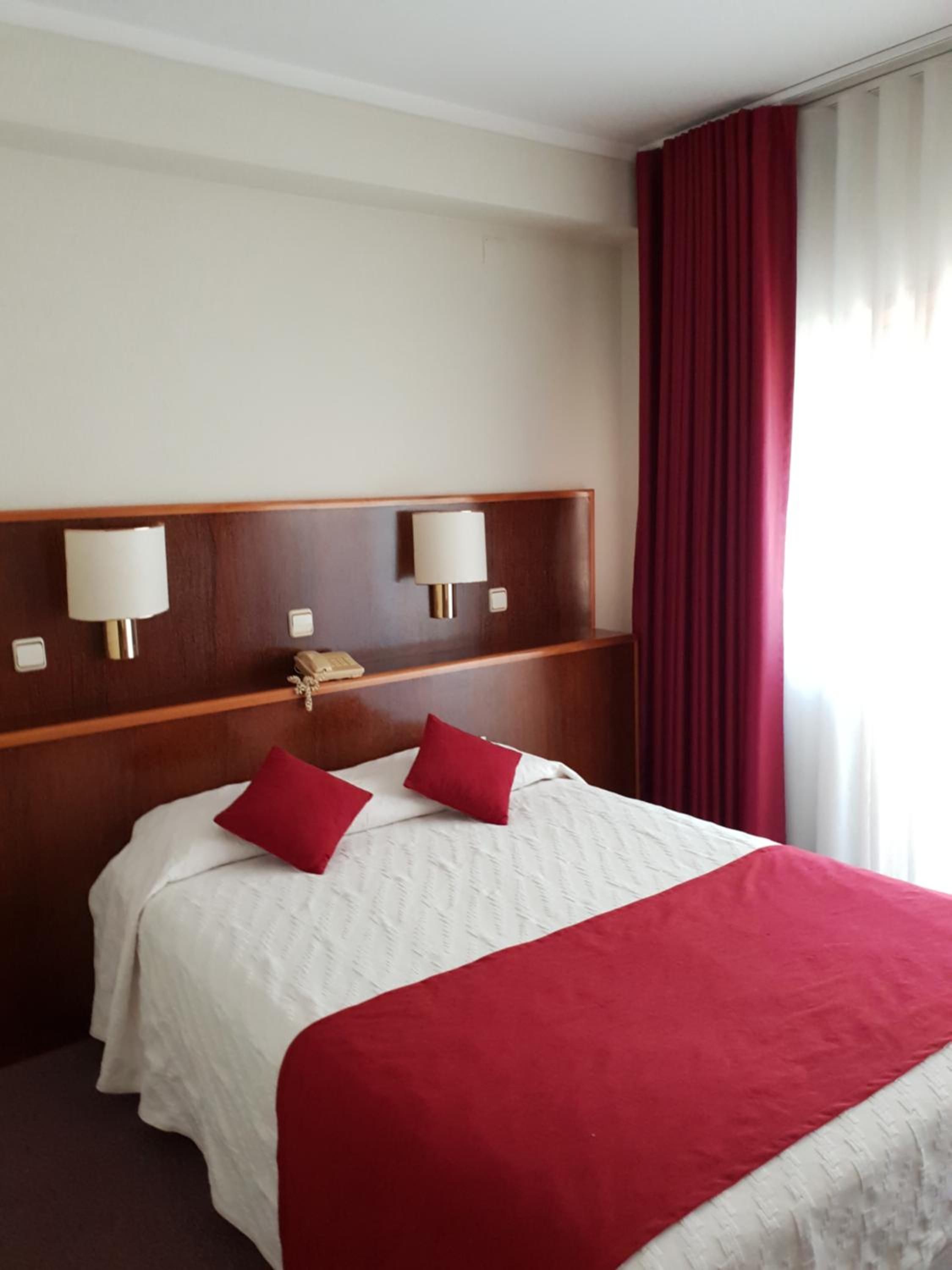 Quarto de hotel com uma cama grande, roupa de cama vermelha e branca, duas luminárias na parede e um telefone na mesa de cabeceira.