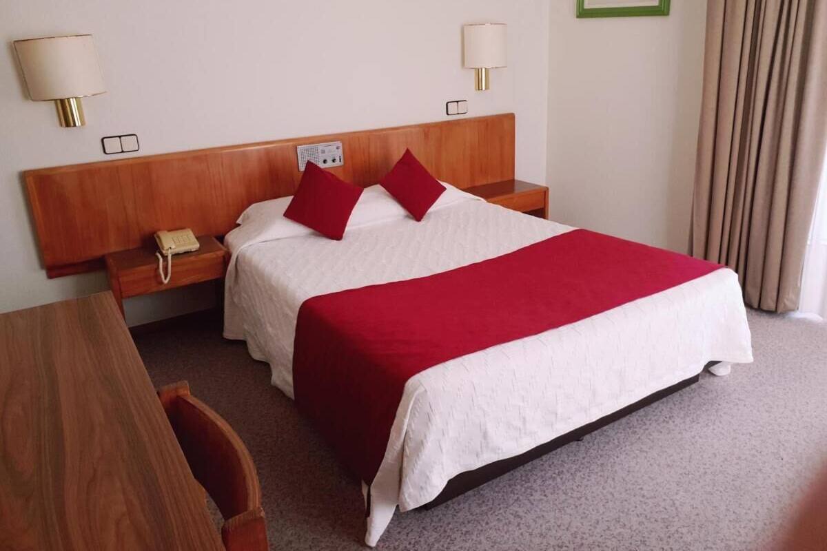 Una habitación de hotel con cabecera de madera, dos mesitas de noche con lámparas, un teléfono, una colcha de cama rojo y blanco, y un cuadro enmarcado en la pared.
