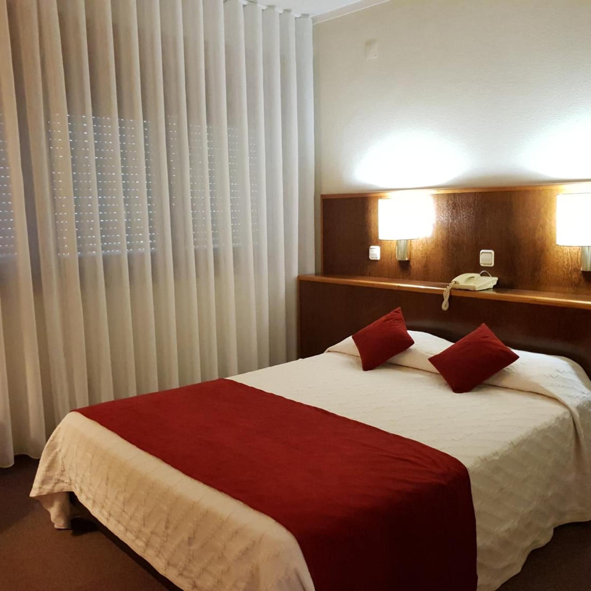 A hotel room with a bed, a bedside table with a lamp, a phone, and a window with blinds.