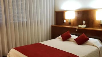 A hotel room with a bed, a bedside table with a lamp, a phone, and a window with blinds.