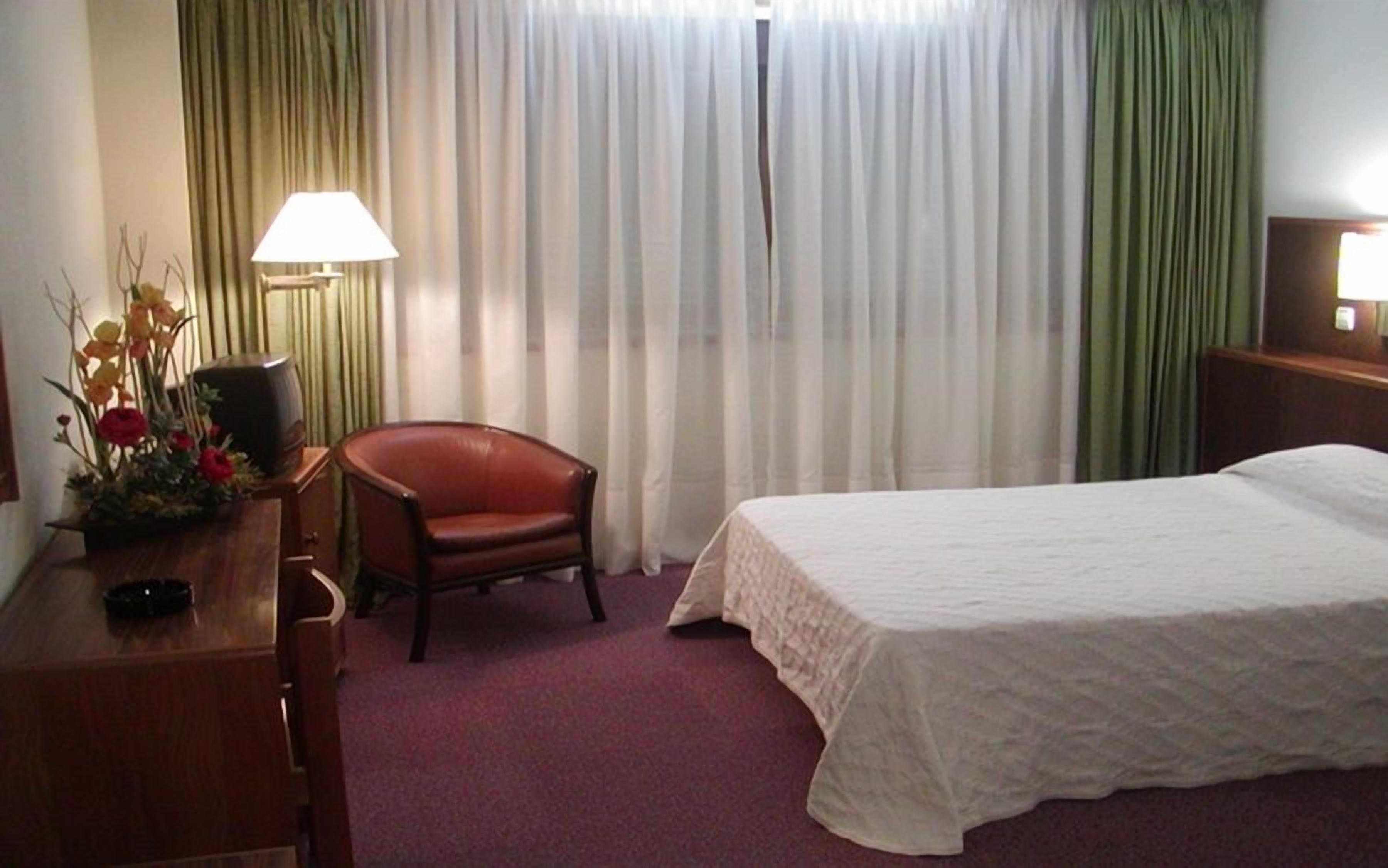 Habitación de hotel con una cama, una silla, un escritorio, una lámpara y una ventana con cortinas.