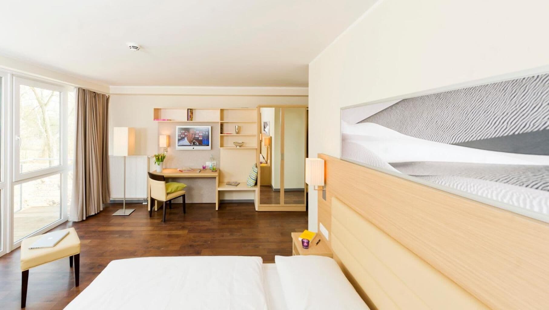 A hotel room with a bed, a desk with a chair, a wardrobe, and a large window.