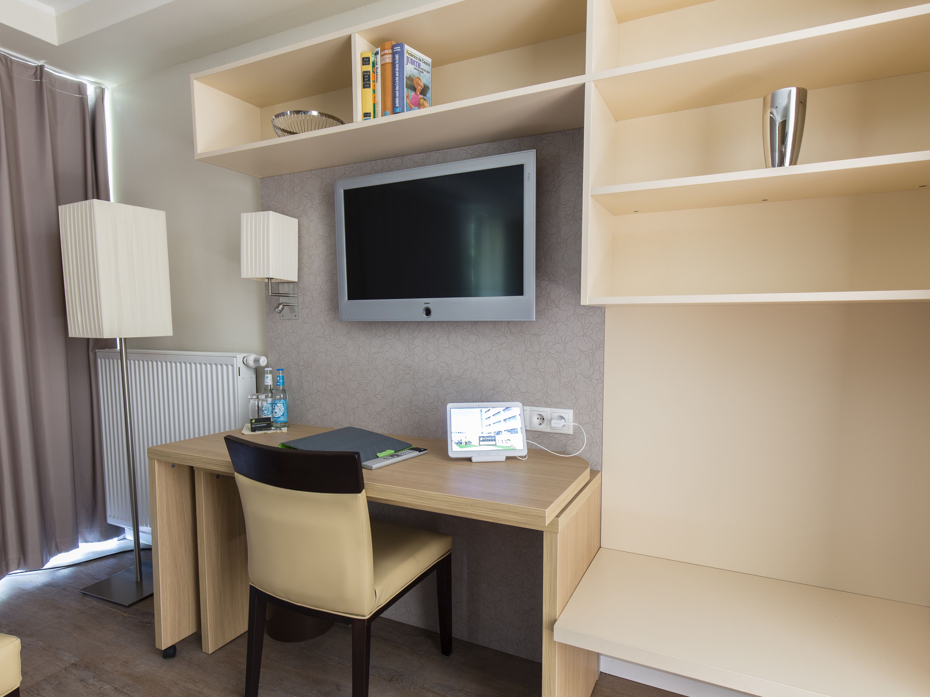Ett hotellrum med ett skrivbord, en stol, en TV, hyllor och ett element.