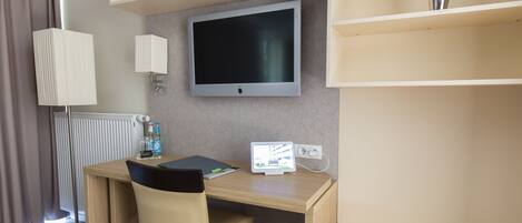A hotel room with a desk, chair, TV, shelves, and a radiator.