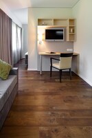 A modern room with a wooden floor, a grey sofa with a green pillow, a desk with a chair, a flat-screen TV, and a lamp.