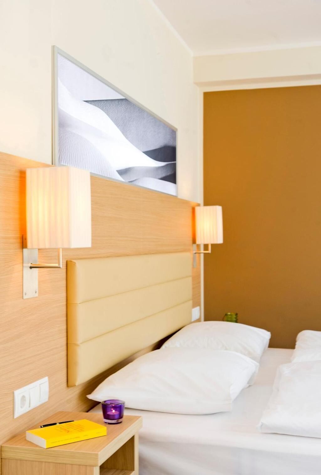 A hotel room with a bed, a bedside table with a lamp, a picture on the wall, and a small nightstand with a book and a candle.