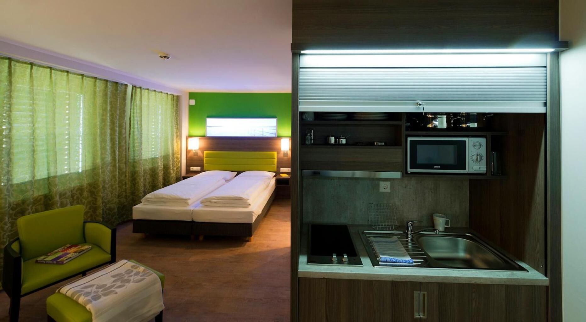 A hotel room with a bed, a chair, a small table, and a kitchenette with a microwave and sink.