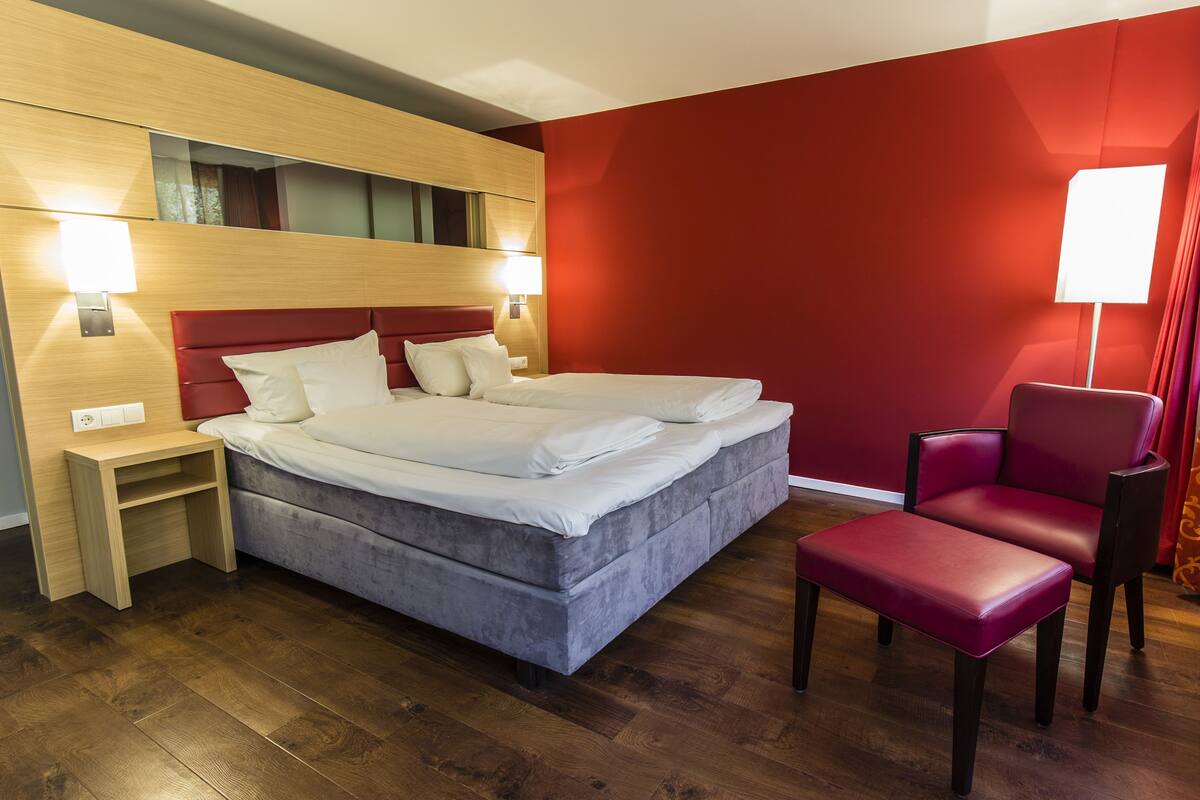Una habitación de hotel moderna con una cama grande, una pared de acento roja y una mesita de noche de madera.