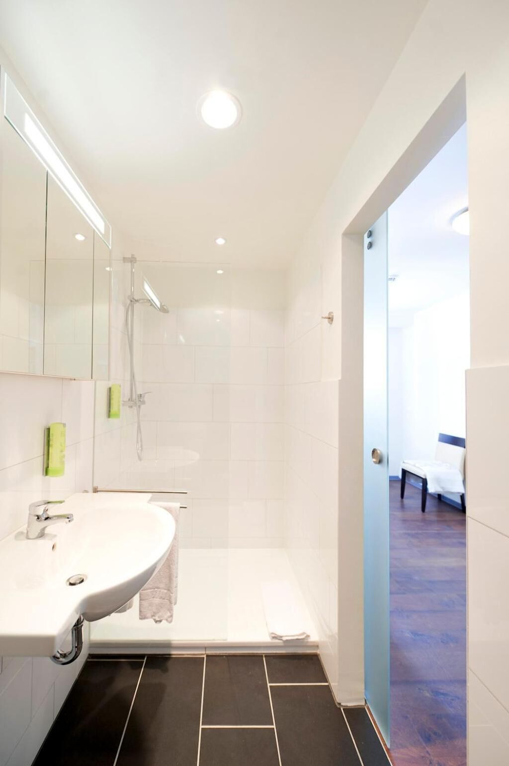 Un baño moderno con un lavamanos grande de color blanco, una ducha y una puerta de cristal que da a un dormitorio.