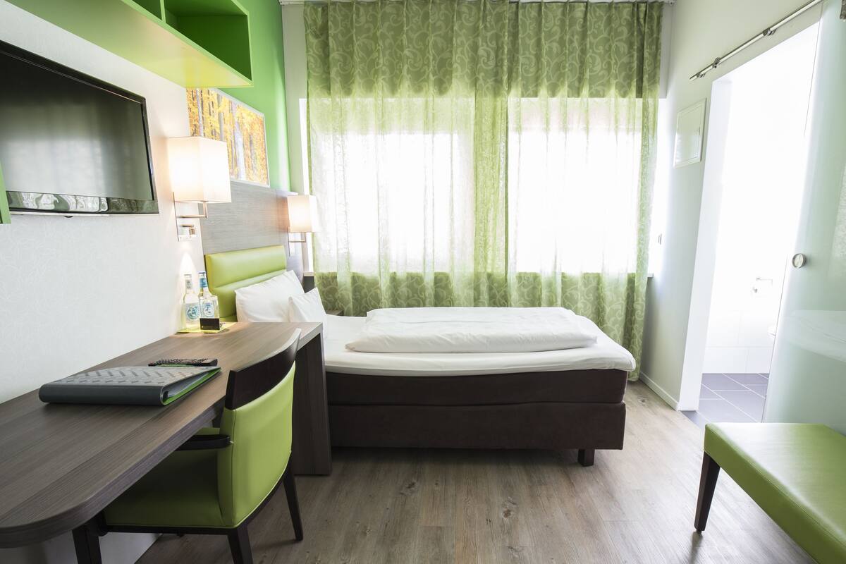 Una habitación de hotel con cama, un escritorio con silla, una lámpara y una ventana con cortinas.